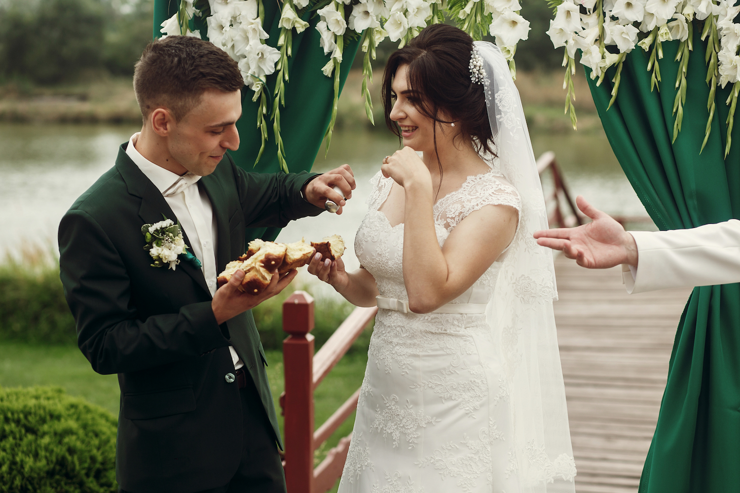 Happy newlywed couple breaking bread at wedding ceremony, handsome groom and smiling bride eating bread near wedding aisle during slavic tradition outdoors